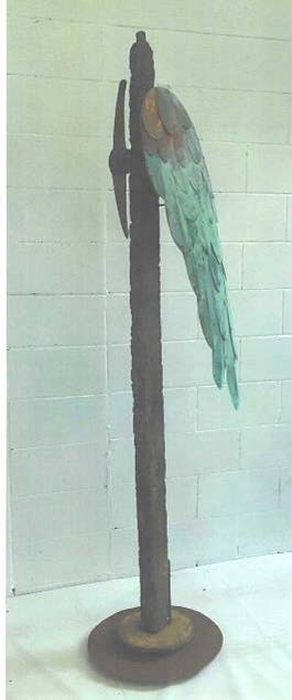 A picture of a rusty metal stand with a pitchfork head on one side and a bird wing on the other. The bird wing is red and gold at the top and cobalt on the bottom.