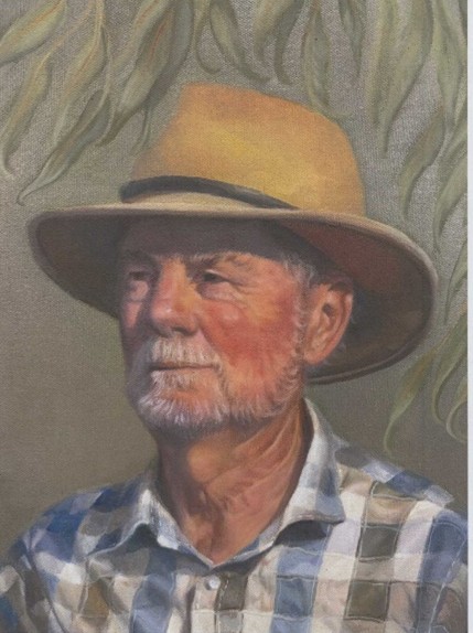 A head and shoulders portrait of a man with a white moustache and beard wearing a simple cotton shirt and a broad brimmed hat, framed by eucalyptus leaves.